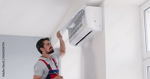Male engineer repairman in work uniform opening air conditioner panel, inspecting condition of AC system after installing, cleaning or repairing. Technician service and maintenance of cooling systems.
