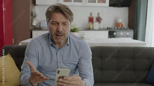 The man gets angry at what he sees on the smartphone.Angry man having problem with cell phone texting. The male user sitting on the sofa at home was frustrated by what the smartphone saw.