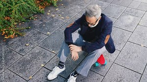 Middle-aged white man with grey hair and beard wearing semi-formal clothes sitting on red skateboard on sidewalk. Cool grandpa. Horizontal outdoor video. High quality 4k footage