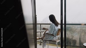Rear view beautiful young woman in dressing gown having coffee at hotel terrace with foggy autumn view slow motion.
