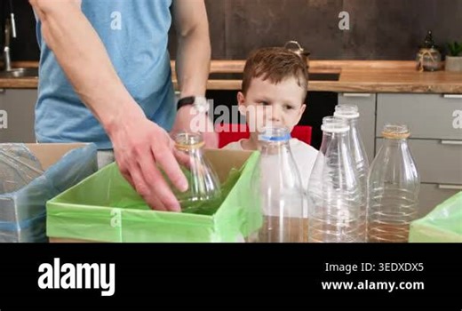 Father and boy sort empty plastic bottles above recycling bins in kitchen. Preschooler boy reaches for clear containers while parent arranges waste for proper recycling