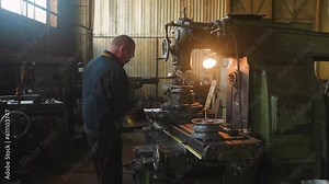 Old worker turns valve of milling machine to fix detail in factory workshop. Man uses vintage lathe equipment to cut out steel material at plant