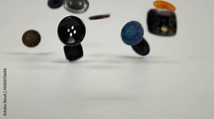 Colorful buttons in different designs and sizes for sewing on clothes fall on a white table, slow motion