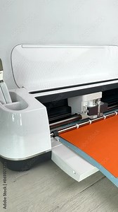 Close up of White Smart Cutting machine home using. Plotter, digital flatbed cut out small details on orange cardboard on blue mat. DIY manual paper crafting project, making gift for family.