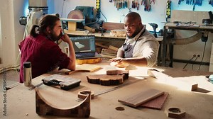 Carpenter looking over blueprints schematics on laptop in CAD software with apprentice. Woodworkers designing custom 3D furniture on notebook in studio, preparing for client order, camera B