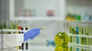 Lab assistant holding green pill, tubes and flask with plant extracts on table