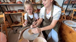 Potter's hands guiding child's hands to work with the pottery wheel. The hands of a woman and a child work on a potter's wheel, make pot from clay. A potter teaches a child pottery, making a clay jug