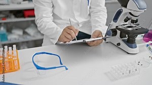 Scientist in lab coat writing notes with microscope and test tubes in a modern laboratory setting.