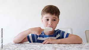 Handsome boy eating popsicle on a stick. He bites off the ice cream with pleasure. Close-up. Child eat in the kitchen or at the dining room table. Tasty dessert. Slow motion. High quality 4k footage