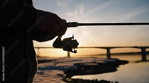 Winter sunset casting golden light on lone fisherman winding fishing line onto spinning rod reel, river bridge reflection mirroring serene landscape