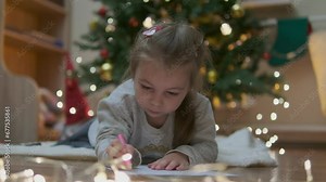 Special orders for Santa. Shot of a beautiful little girl smiling while writing a letter lying near the presents under the Christmas tree copyspace