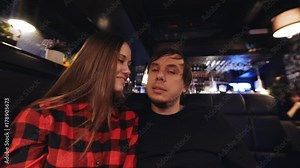 Young attractive couple is having a good time in a cafe: looking into the camera, lady is smiling, man is kissing her on a cheek and speaking to someone behind the camera. Camera is zooming in