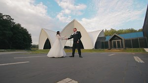 Bride and Groom Dancing Outdoors by Modern Architecture. A bride and groom dressed in formal wedding attire are dancing outdoors in front of a modern architectural building with a unique design.