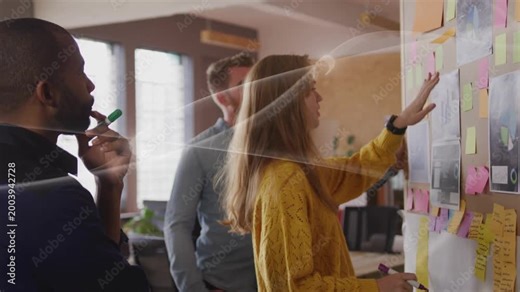 Woman in sweater leaning, scanning sticky notes on board, organizing project while men watching