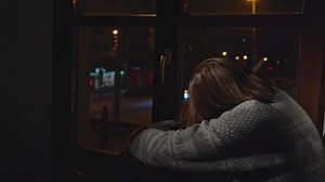 Lonely young woman looks out night window. Media. Young woman sits on window and looks sadly at night city. Woman with depression sits alone on window of house