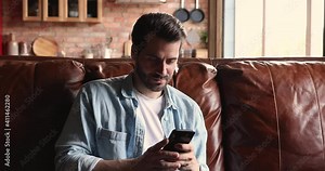 Handsome young man sit on leather sofa at home using smartphone spend leisure time online, chatting buying on internet, enjoy comfort modern tech usage, speed wi-fi connection satisfied client concept