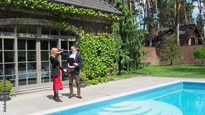 Property manager and female customer stand by big swimming pool of summer cottage residence. Businesswoman in red dress looks at cottage yard