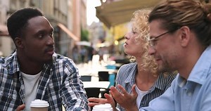Three diverse good looking happy people sitting at table in cafe terrace and sipping coffee while enjoying summer day. Friends drinking coffee in morning in center city and talking