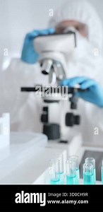 Close up of test tubes with blue liquid opposite a scientific researcher wearing protective gear tuning microscope lenses in laboratory. Medicine, healthcare and science concept Stock Video Footage - Alamy