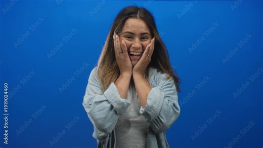 Woman clapping hands with open mouth and hands on cheeks in studio blue backdrop; surprise celebration.