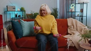 Mature woman doing dumbbells weight lifting fitness exercises at home apartment indoors. Senior old grandmother workout training strength practicing effort in living room on couch. Sport motivation