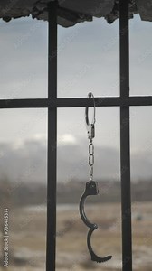 Vertical screen: Hanging steel police handcuffs are undone on the bars of a prison cell window overlooking the landscape. Prisoner release concept. The convict is free after punishment. Atonement