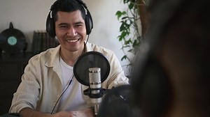 Businessman giving interview and laughing, sitting at table while broadcasting online on radio. Close-up view of young handsome man speaks and looks with smile, then laughs and sits at desk
