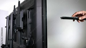 Closeup isolated shot of a man's hand holding and pressing a remote control while facing a TV, with the main subject view being the rear of the television, which is attached to a metal stand.