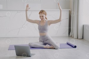 Slim sporty girl stretching on mat. Online remote fitness classes student watching video on laptop.