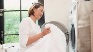 slow motion scene on young asian woman trying to push blanket fabric into washing machine to washing after unpleasant smell in utility room for routine lifestyle and housework concept