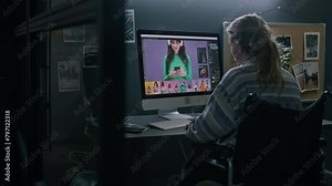 Medium rear of blonde female graphic designer with disability sitting in wheelchair at desk in front of computer monitor, retouching and editing colourful commercial images, alone in dark studio