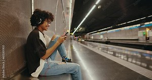 Beautiful African-American girl sitting in the subway, listening to music, chatting on her smartphone and waiting for her train. A young black woman is waiting for her train in the subway.