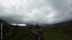 A white man wearing FPV goggles and a down jacket sits in the mountains and controls an FPV acrodrone quadcopter flying around him.