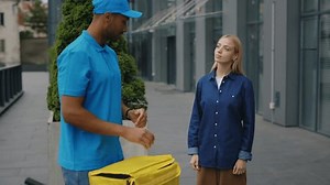 Positive African American Postman in Blue Uniform Delivering Food in Paper Packages to Beautiful Customer Young Woman in Modern Business District. Work Order Delivery, Transportation