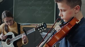 Two girls playing guitars and boy with violin practicing song together during music class in school