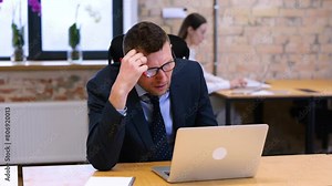 Realtime video. Caucasian man is sitting and looking at his computer in office with bricks walls, male takes off his glasses and looks exhausted.