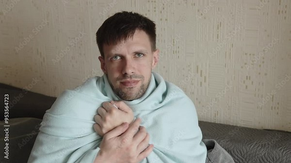 Caucasian man shivering and huddled in blanket at home. Chills from high temperature and portrait of person trying to warm up in cold weather during illness. Energy crisis.