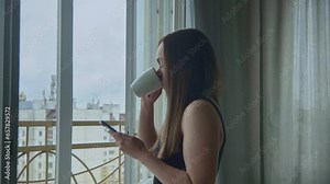 Confident brunette woman coming to open window with the great city view, drinking hot tea or coffee and scrolling screen of her smartphone, checking messengers and social media. Mid shot, side view