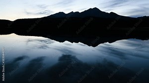 Time lapse of a late evening mountain setting with sharp mountain peaks and clouds reflecting in a large calm lake.