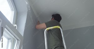 man standing on ladder and attaching ribbon tape on wall. window is on the left, it is daytime.