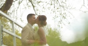 A bride in a simple gown and groom in a beige suit kiss by a white country fence under a tree as seen from behind grass in the farm field