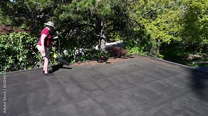 Middle aged woman with a cordless leaf blower cleaning tree debris off a flat roof in the spring