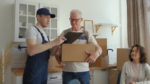 Courier brings cardboard box to new apartment of senior couple asking to sign paper of conformation. Elderly grey-haired man in glasses receives box with things