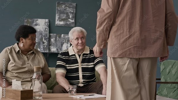 Stab shot of Caucasian female family psychologist adjusting and setting up camera at beginning of therapy session with multiethnic senior couple undergoing marriage crisis