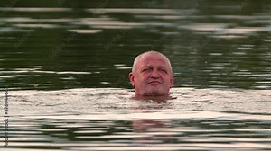 The man stuck his hairless head out of the water and swims. Close up of a man's face. He moves his arms and stays on the surface. Soft daylight. The surface of the reservoir has a greenish tint.