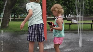 Little happy children playing with water at splash pad in the local public park on hot sunny summer day. Small beautiful kids having fun at fountain playground.