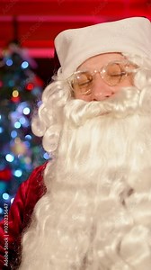 Santa Claus smiling on camera indoors. Portrait of a real Santa with white beard, in glasses and red costume standing in the living room on Christmas tree background. Vertical video