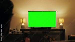 Person watching television with chroma green screen. Unrecognizable female person switching channels on tv with remote controller, sitting on sofa in living room.