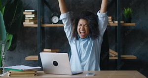 Excited young african american business woman enjoying winning sits at table with laptop computer working in modern office. She gets great news and is delighted. Showing yes gesture