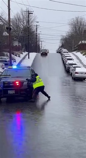 Officer Slips on Black Ice as Cars Slide Down Hill 🚓 Residential City Street — Winter 2025 Vertical smartphone footage shows a steep residential street coated in reflective black ice as a police cruiser sits below with lights flashing. An officer attempts to cross the road, loses footing, and falls just moments before vehicles begin sliding uncontrollably downhill. Several cars crash into a line of parked vehicles, shoving them onto the snowy sidewalk as bystanders shout warnings. The ice refle